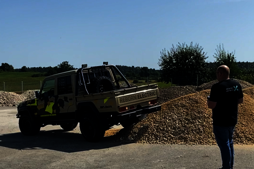 A Toyota Landcruiser with double cab for underground mining in brown with black and neon-colored highlights, drives with the right rear wheel on a pile of gravel, controlled by an Arnold NextG employee who looks at the Toyota and has a remote control in his hand.