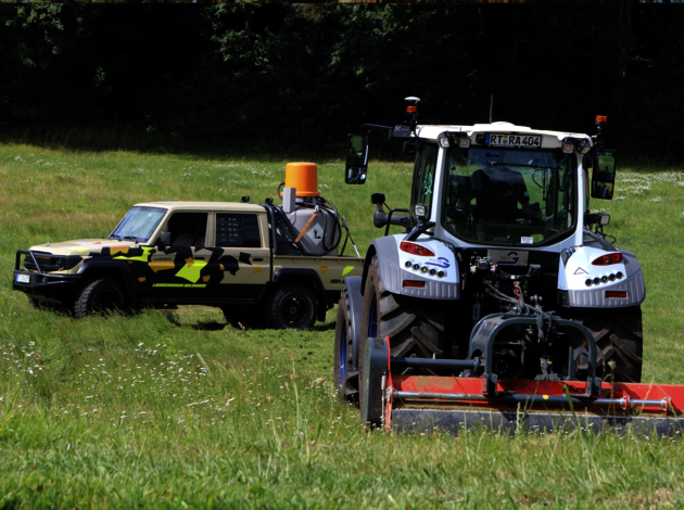 Autonomous tractor with mounted implement and modified off-road vehicle equipped with sensor technology during field testing on a grassy meadow.