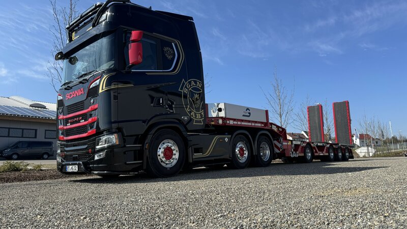 A black and red Scania truck with a flatbed trailer attached is parked on gravel in sunny weather. 