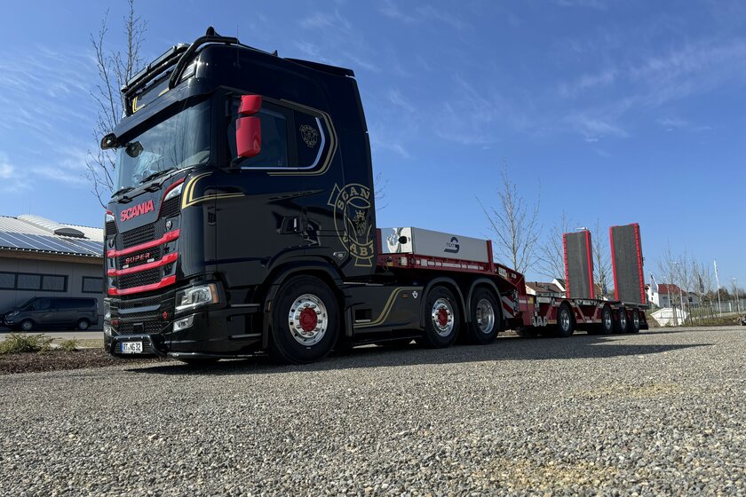 A black and red Scania truck with a flatbed trailer attached is parked on gravel in sunny weather. 