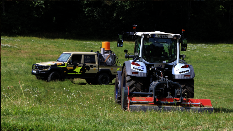 A tractor with a mower is driving across a meadow, followed by an autonomous Land Cruiser with camouflage paintwork and a fuel tank unit on the loading area. The edge of a forest can be seen in the background. The scene shows the use of modern drive-by-wire technology in agricultural engineering.