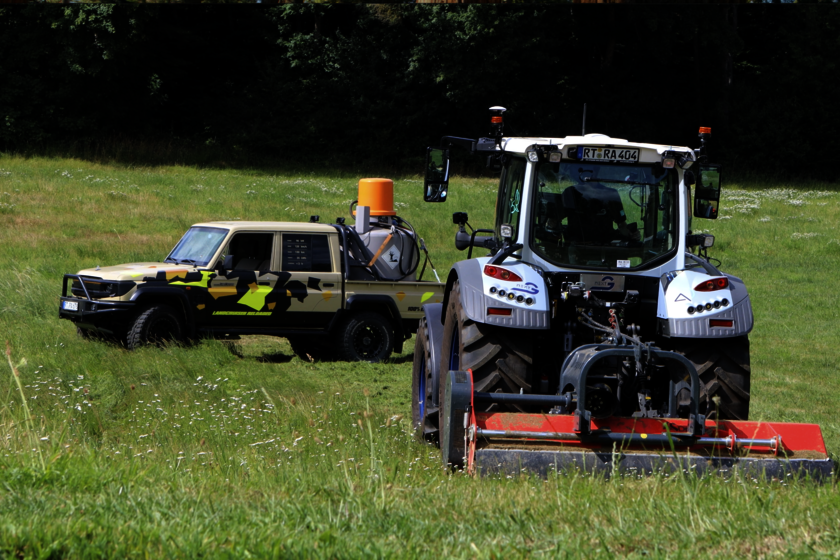 A tractor with a mower is driving across a meadow, followed by an autonomous Land Cruiser with camouflage paintwork and a fuel tank unit on the loading area. The edge of a forest can be seen in the background. The scene shows the use of modern drive-by-wire technology in agricultural engineering.