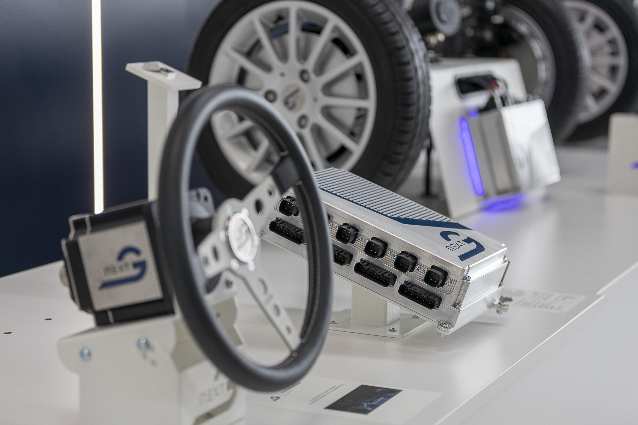 On a table are several objects arranged diagonally across the image. In the lower left corner is a steering wheel with a motor attached to the back; in the center is an NX NextMotion central control unit; and in the background is a platooning system consisting of two vehicle axles that can be steered using a joystick.