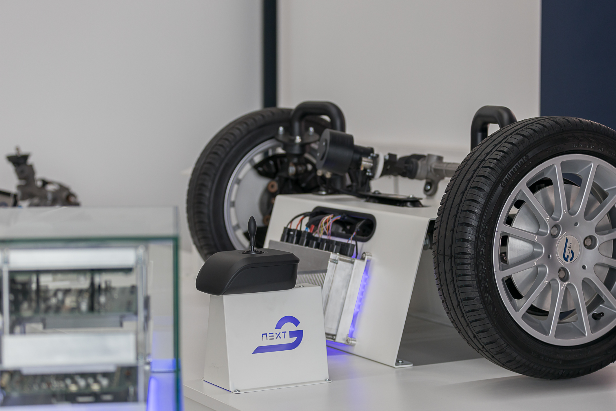 In the lower left corner, you can see a blurred glimpse of the NX NextMotion exhibit. In the background is the front axle, along with the wheels of a car, which is connected via cables to an NX NextMotion control unit that can be operated left and right using a joystick positioned in front of it.