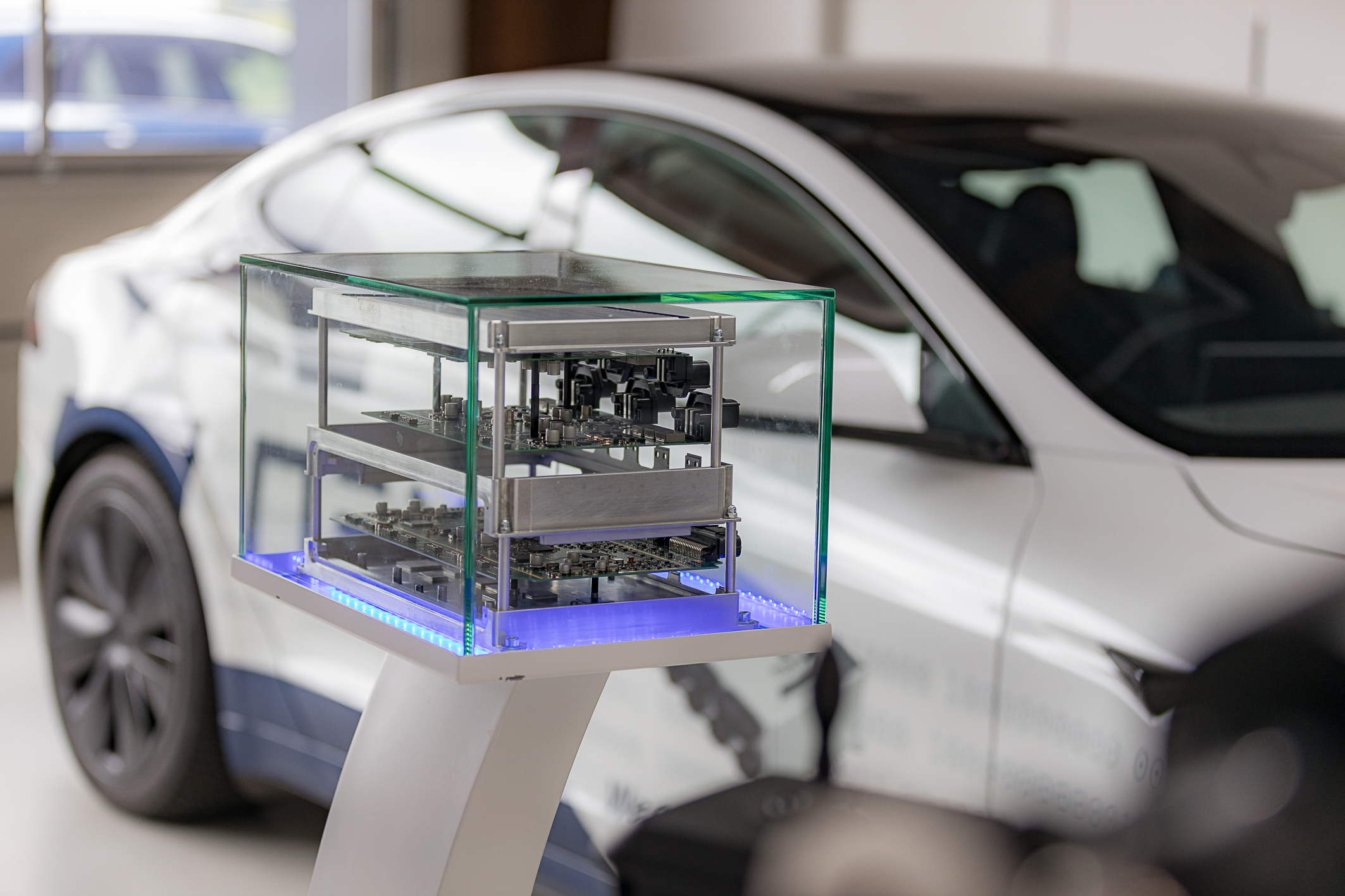 In the foreground is the NX NextMotion, which has been pulled apart and arranged on three levels so that the circuit boards inside are visible. The entire display is presented in a glass showcase. In the background, a white Tesla is visible out of focus. 