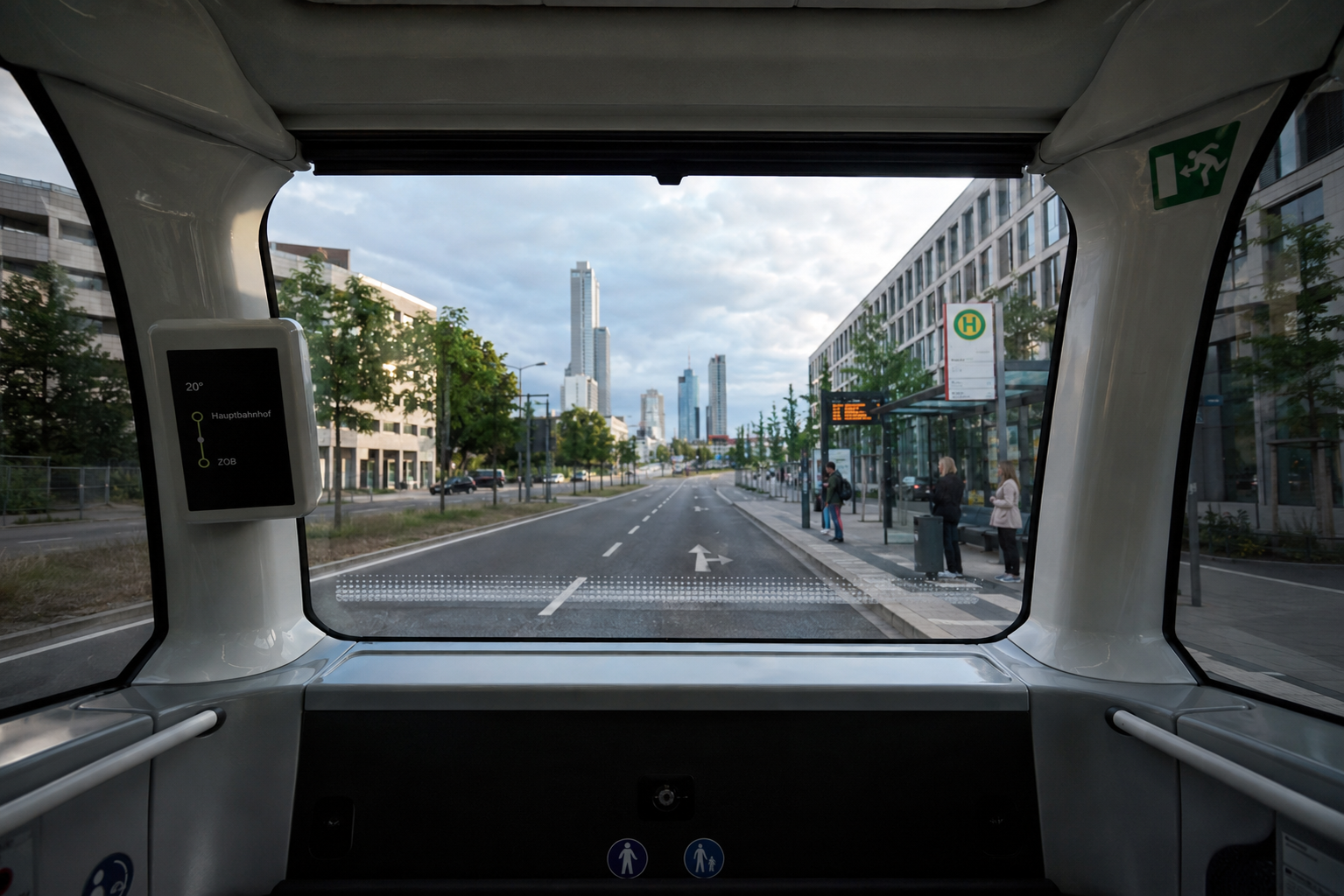 The view through the windshield of an autonomous shuttle bus traveling in the left lane. On the right is a bus stop where several people are already waiting. In the background, a large city with skyscrapers is visible.