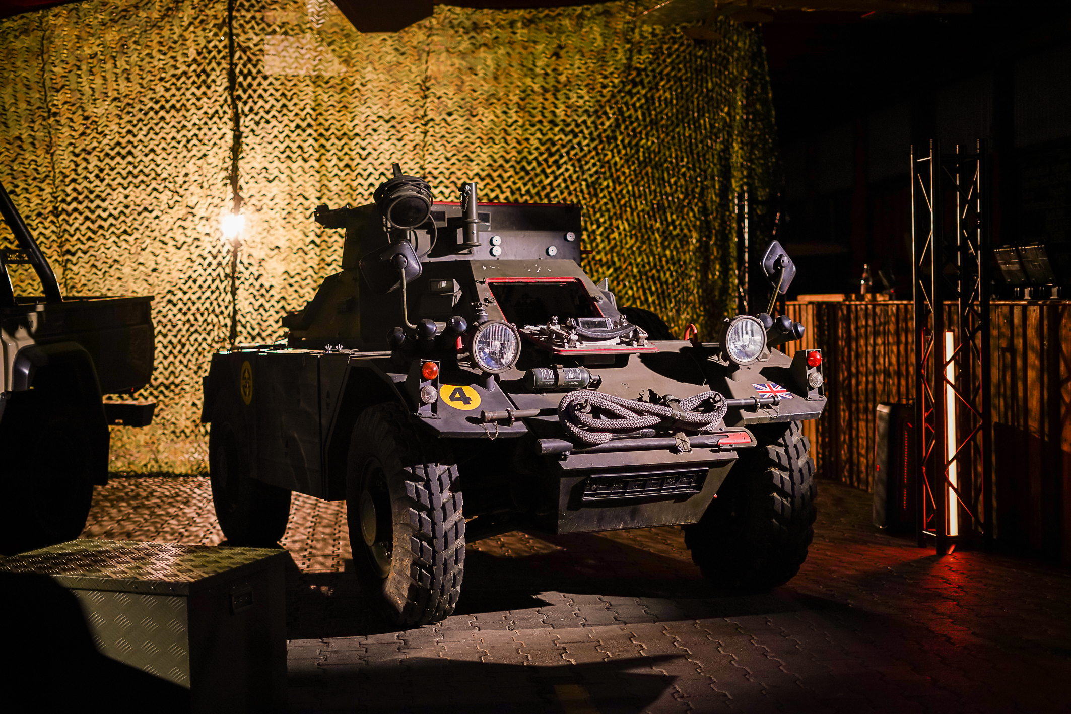 An older, small military vehicle stands on a paved stone floor with a camouflage net in the background. The image is rendered in shades of green and dark yellow. 