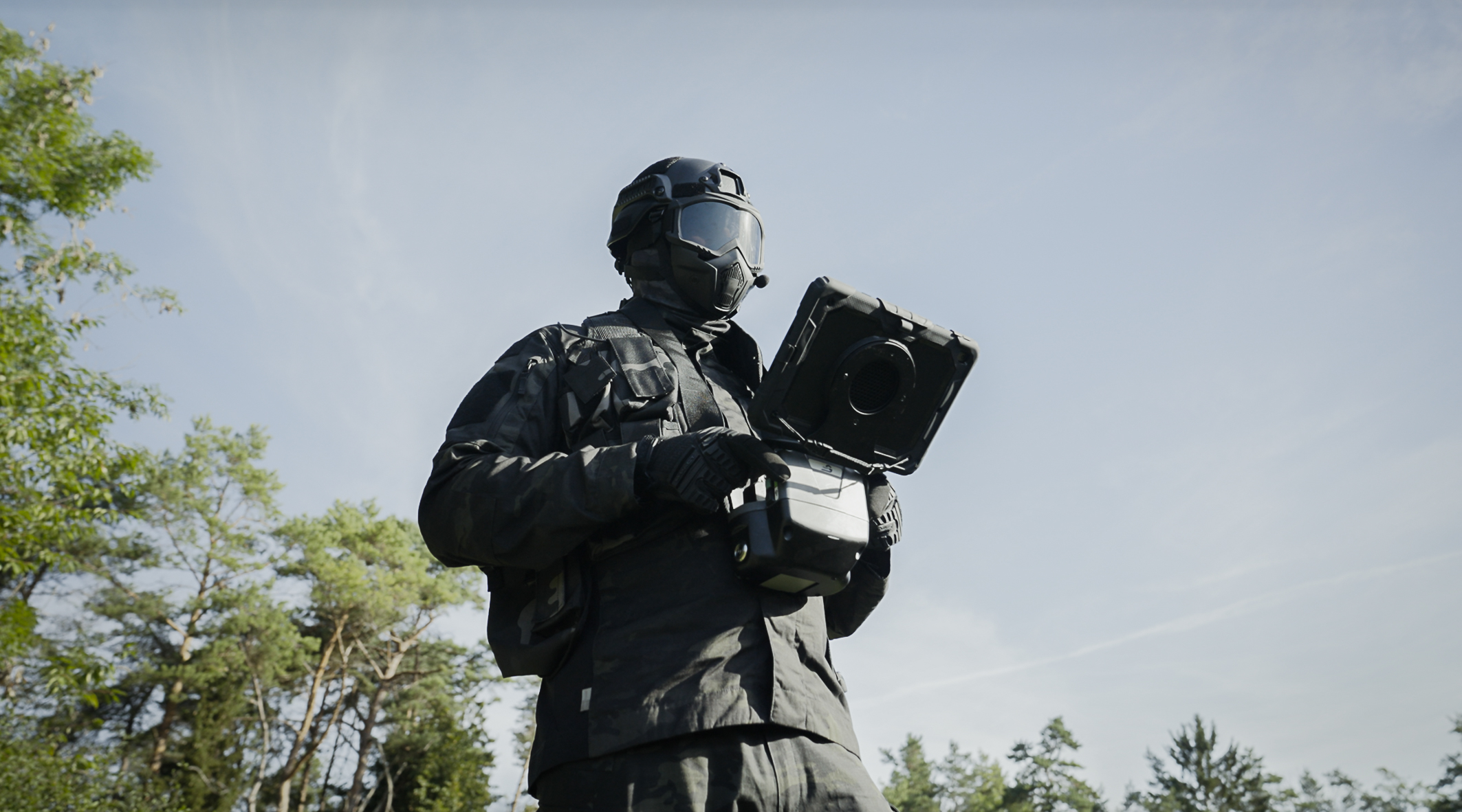 A person is standing at the edge of a forest under a blue sky. The person is dressed in camouflage and is wearing a gas mask. In his hands, he is holding a remote control with an iPad attached to the top.