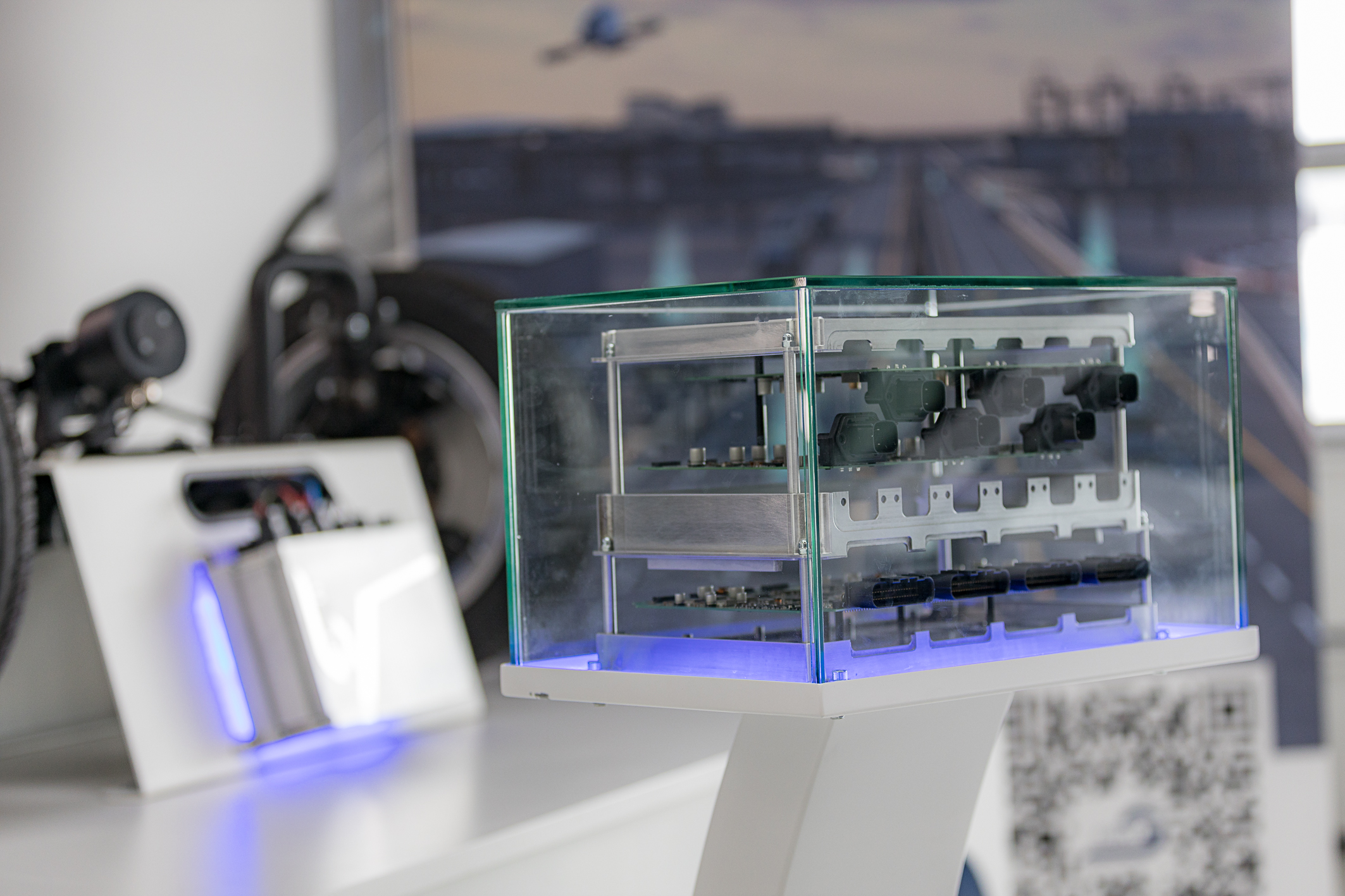 Behind a small glass display case containing an exhibit is an open NX NextMotion unit arranged in tiers. In the background, another exhibit—an axis also equipped with an NX NextMotion—can be seen out of focus.