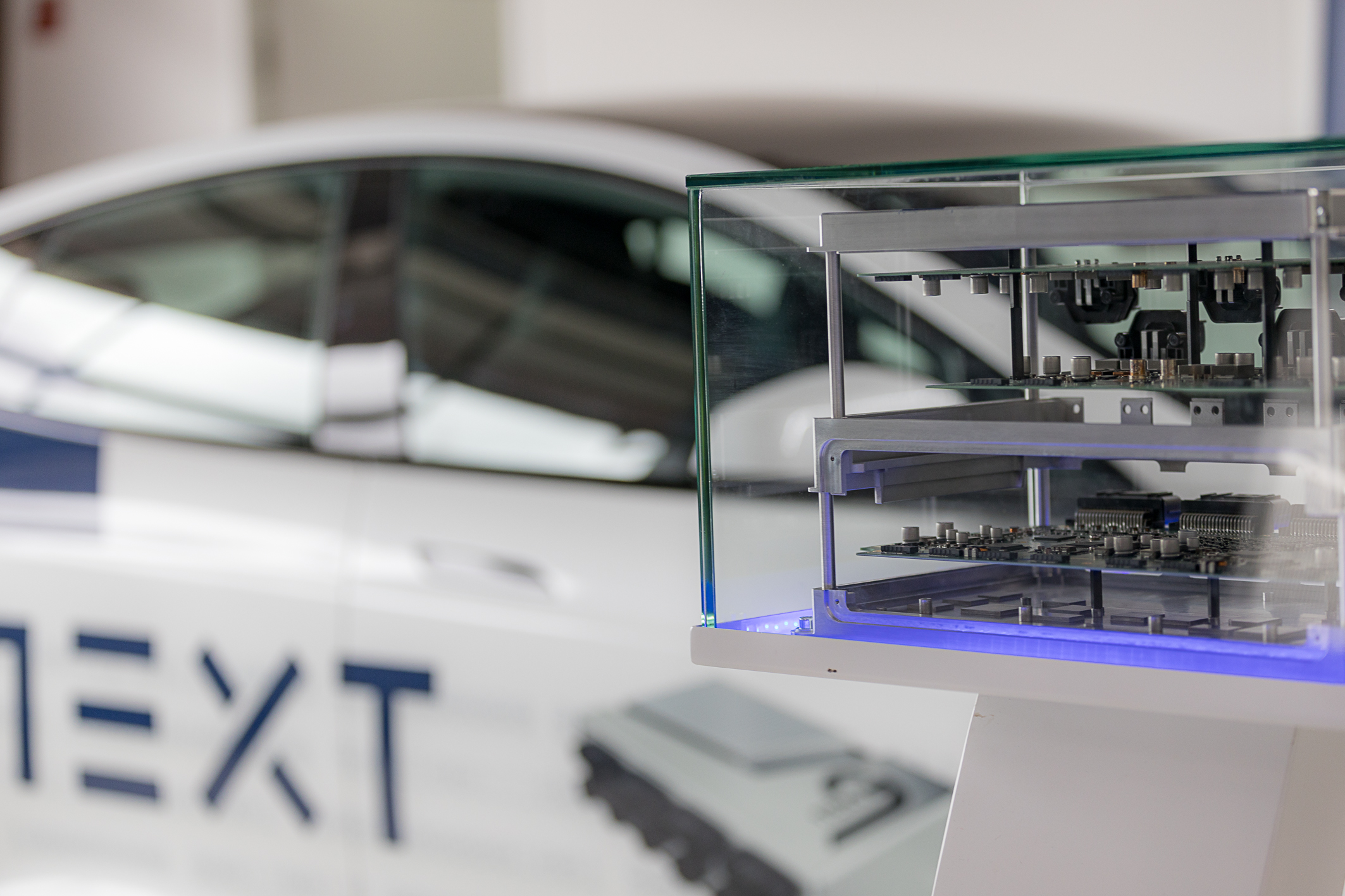 Behind a glass display case, which houses an exhibit, there is an open NX NextMotion unit arranged in tiers on the right-hand side. In the background on the left, a white Tesla with NextG decals can be seen out of focus.